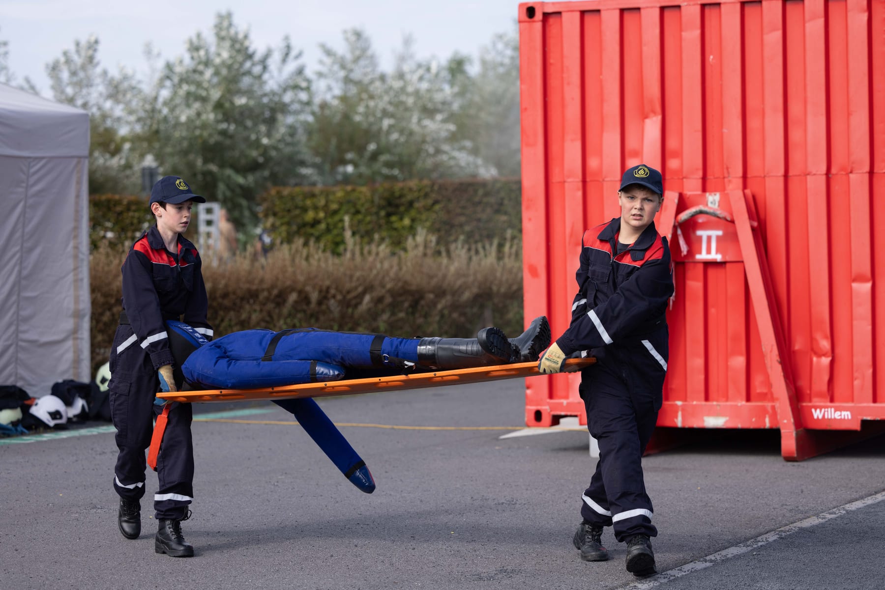 Twee jeugdbrandweerleden dragen een oefenpop op een brancard weg van een container